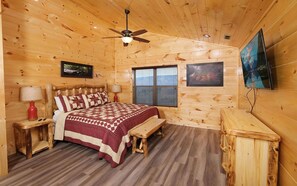 A wooden cabin bedroom with a bed, two bedside tables, a bench, a ceiling fan, a window, and a TV mounted on the wall.