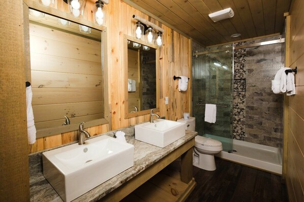 A wooden bathroom with two sinks, a toilet, and a glass shower enclosure.