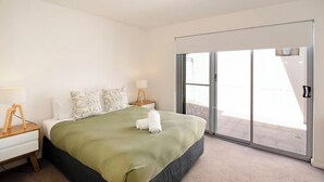 Une chambre à coucher comprenant un lit, une table de chevet, une lampe, une fenêtre et une porte-fenêtre coulissante donnant sur un balcon.