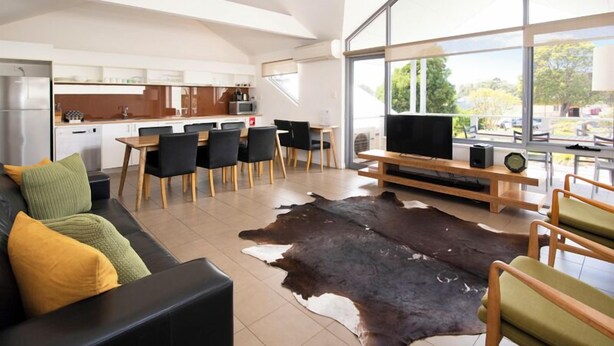 Un salon moderne avec un canapé en cuir, un coin repas comprenant une table et des chaises en bois, et une grande fenêtre donnant sur l’extérieur.