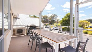 Une terrasse extérieure avec une table et des chaises en bois, un barbecue et une vue sur un parking et des arbres.