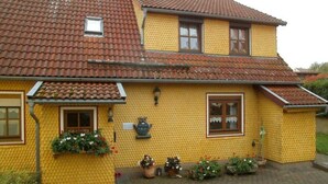 Een twee verdiepingen tellend huis met een gele gevel en een dak bedekt met rode dakpannen. Op de begane grond zijn er twee ramen en op de bovenverdieping één raam. Een pad van baksteen leidt naar de ingang.