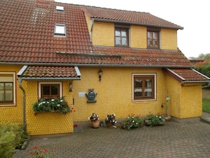 A two-story house with a yellow exterior and red-tiled roof. There are two windows on the ground floor and one on the upper floor. The house has a brick pathway leading to the entrance. - Vacation apartment Landidylle (Birx)