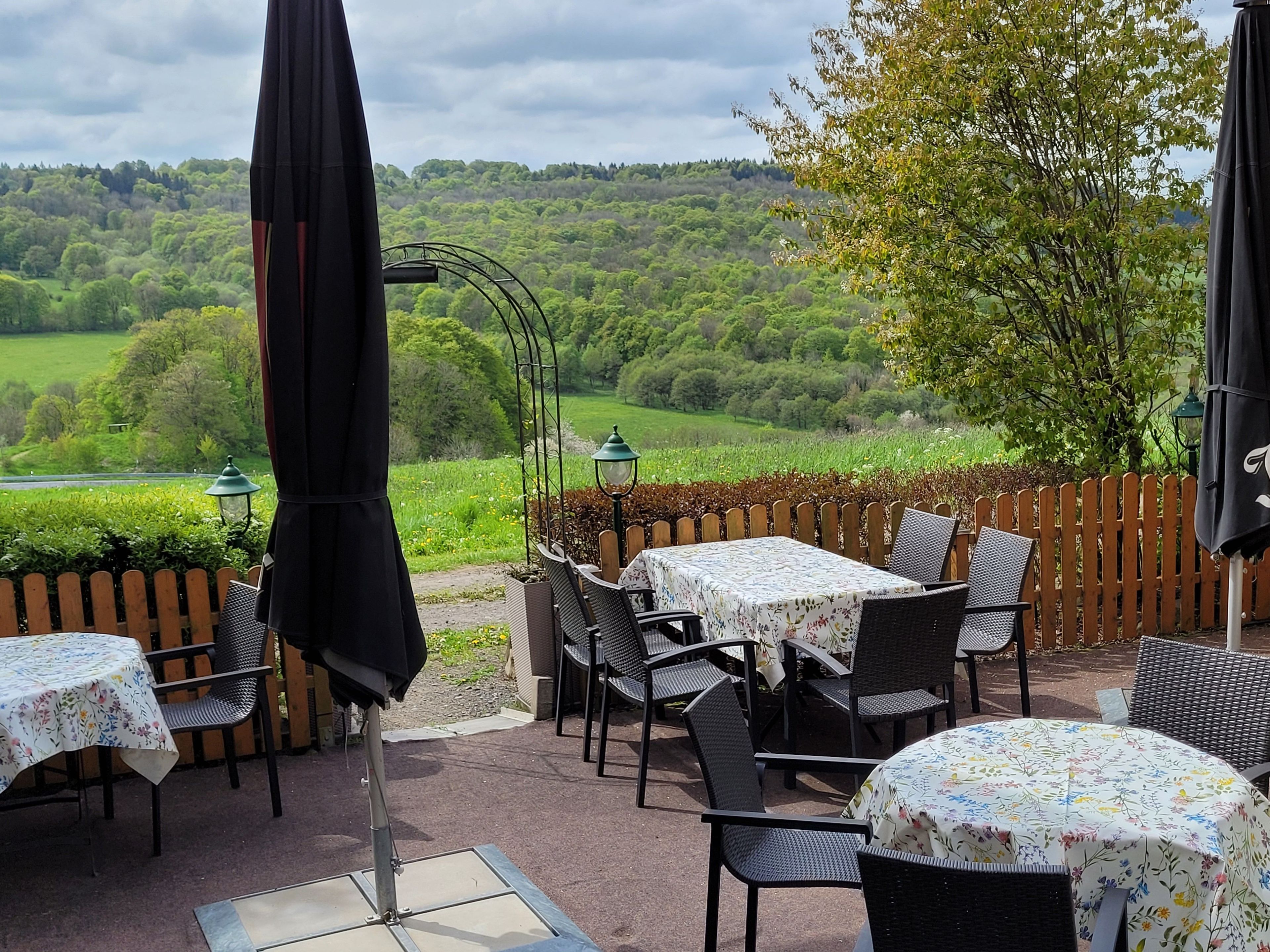 Une terrasse extérieure avec des tables et des chaises, entourée d’une clôture en bois, donnant sur un paysage verdoyant et luxuriant.
