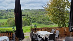Een terras buiten met tafels en stoelen, omringd door een houten hek, met uitzicht op een weelderig groen landschap.