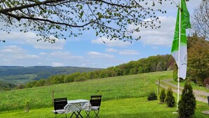 Een gazon met een tafel en stoelen, een vlaggenmast met een vlag, en een mooi uitzicht op heuvels en bomen.