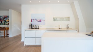 Eine moderne Küche mit weißen Schränken, einer Edelstahl-Spüle und einer Kaffeemaschine. Ein Essbereich mit Holztisch und Stühlen ist ebenfalls vorhanden.