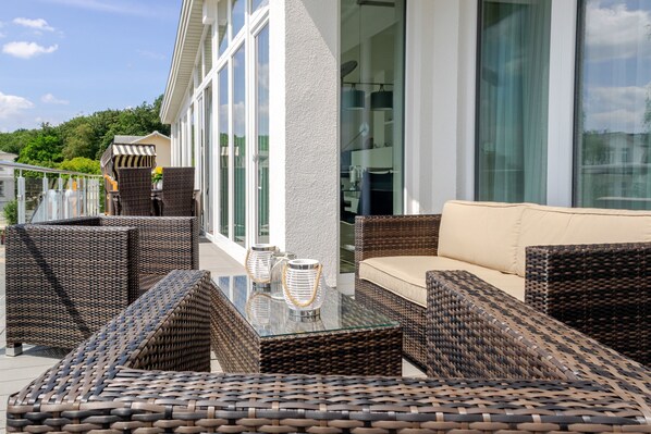 An outdoor seating area with wicker furniture and a glass-top table, overlooking a modern building with large windows.