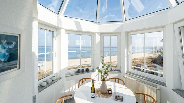 Una habitación soleada con un tragaluz grande, una mesa redonda con sillas, un jarrón con flores y vistas a la playa.