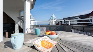 Una mesa de madera con una taza azul, un bol de fruta y un menú en un balcón con vistas a un puente.