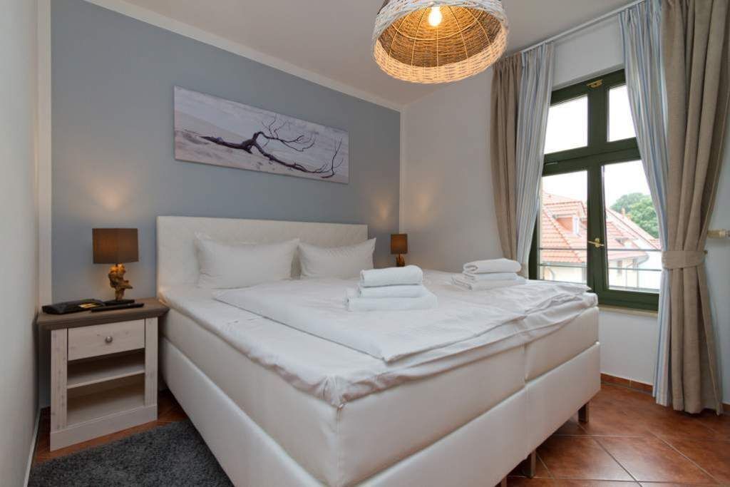 A neatly made bed with white linens, a bedside table with a lamp, a wall-mounted artwork, and a window with curtains.