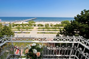 Ein Balkon mit Metallstühlen und einem Tisch, der Blick auf einen Strand mit einem Steg und einen klaren Himmel bietet.