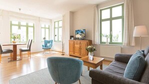 Un salon bien éclairé, comprenant un coin repas, de grandes fenêtres et un téléviseur posé sur un meuble en bois.