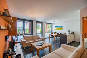 Una sala de estar moderna con sofá, mesa de centro y comedor. Amplias ventanas ofrecen luz natural y vistas al follaje exterior.