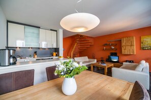 Una cocina moderna con armarios blancos, una escalera de madera, un comedor con mesa de madera y una sala de estar con sofá blanco y televisor.