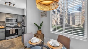 A modern kitchen with stainless steel appliances, a dining area with a glass table and woven placemats, and a large window with blinds.