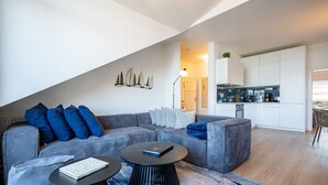 A modern living room with a grey sofa, two black coffee tables, and a blue rug. A kitchen is visible in the background. - Villa Linquenda, App. 6 (Heringsdorf)
