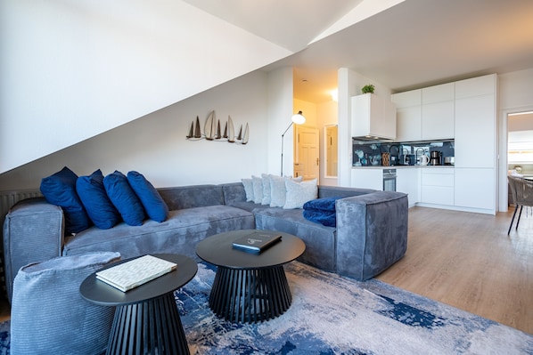 Un salon moderne avec un canapé gris, deux tables basses noires et un tapis bleu. On aperçoit une cuisine au fond.