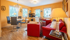 A modern living room with a red sofa, a dining table with chairs, a chandelier, and a kitchen in the background. - Inselstrand, App. 314 (Heringsdorf)