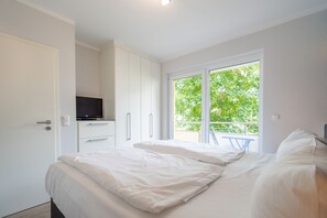 Un dormitorio con cama, televisión, puerta corredera a un balcón y armario blanco.