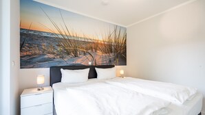 Ett sovrum med en stor säng, vita sängkläder, två sängbord med lampor och en stor väggmålning som föreställer en strandscen.