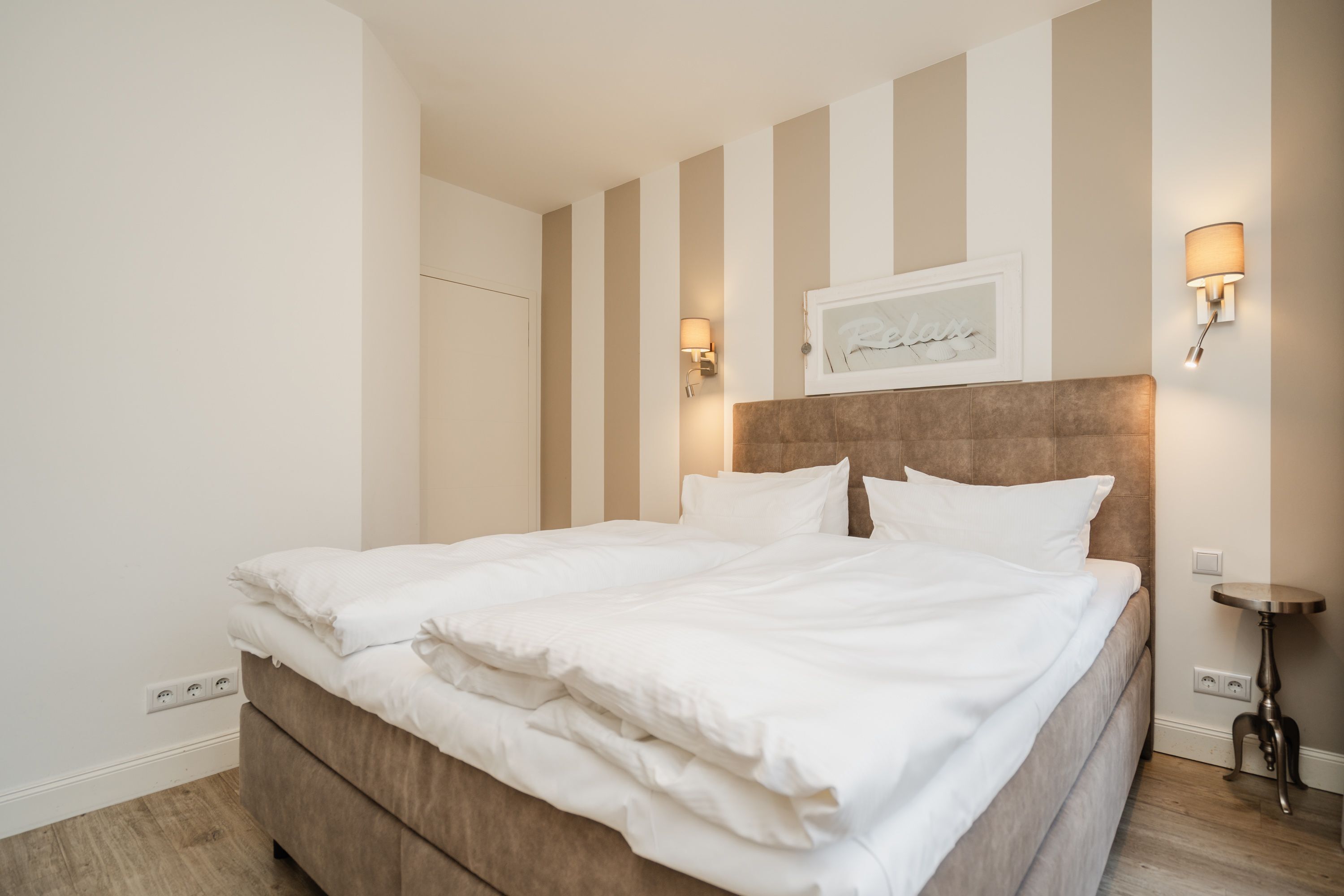 A neatly made bed with white linens in a room with striped walls and a headboard. There are two wall-mounted lamps on either side of the bed.