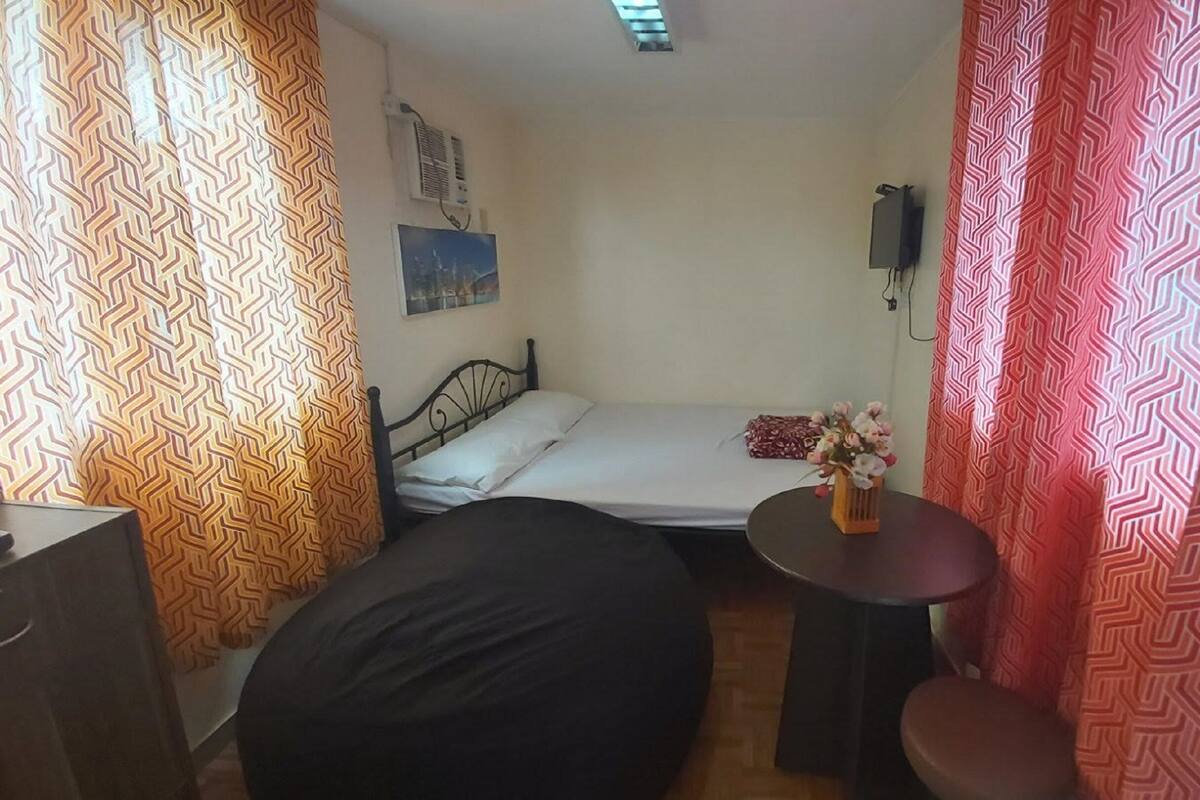 Une petite pièce, de taille modeste, comprenant un lit simple, une petite table et un fauteuil en forme de poire. On y trouve également un téléviseur fixé au mur et une fenêtre avec des rideaux.