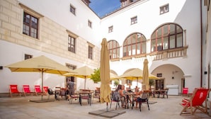 An outdoor café area with tables and chairs, umbrellas, and a building with arched windows and a balcony. - Chateau Třešť (Trest)
