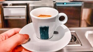 A hand holding a white cup with a blue design, in a kitchen setting with espresso machines and shelves of bottles. - Chateau Třešť (Trest)
