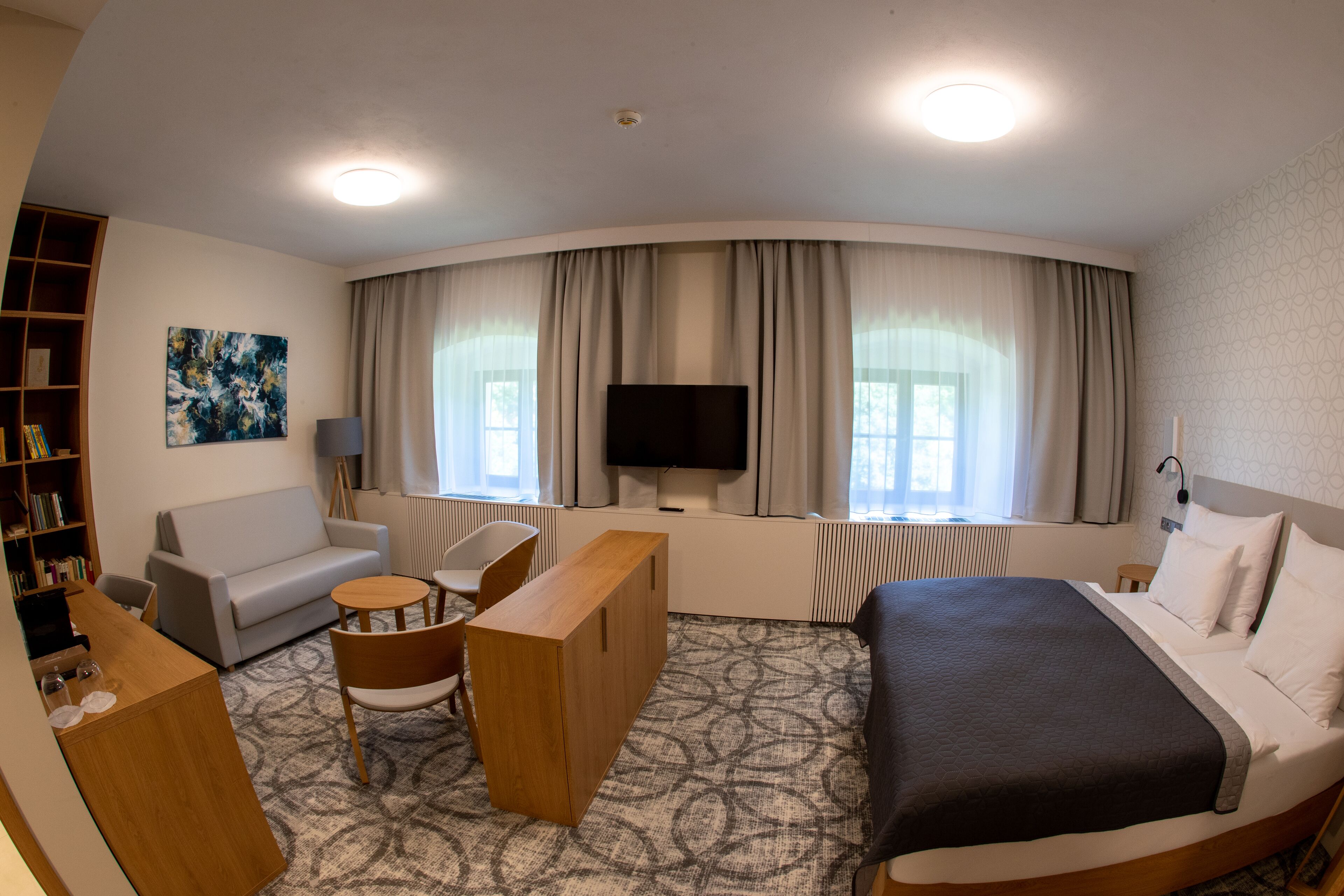 A hotel room with a bed, a sofa, a desk, a chair, a TV, and a bookshelf.