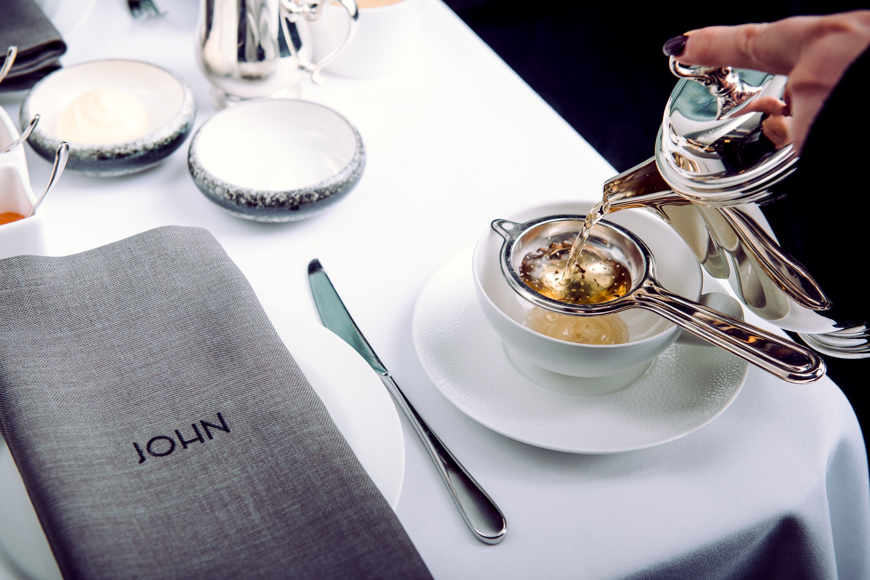 A table setting with a cup of tea, a spoon, and a napkin with the name "JOHN" on it.