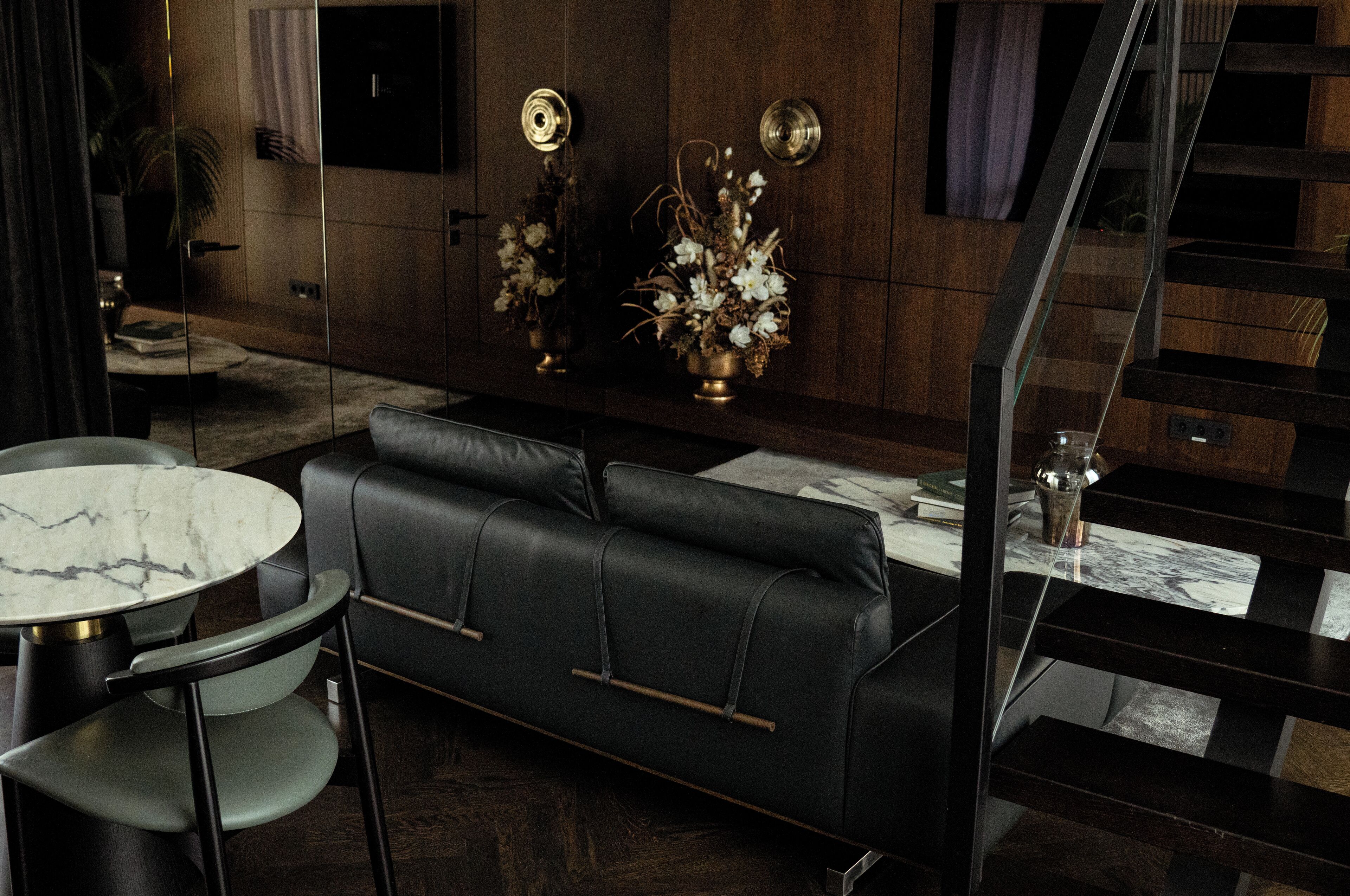 A modern living room with a black leather sofa, a marble coffee table, and a glass-top dining table. There is a staircase leading to an upper level.