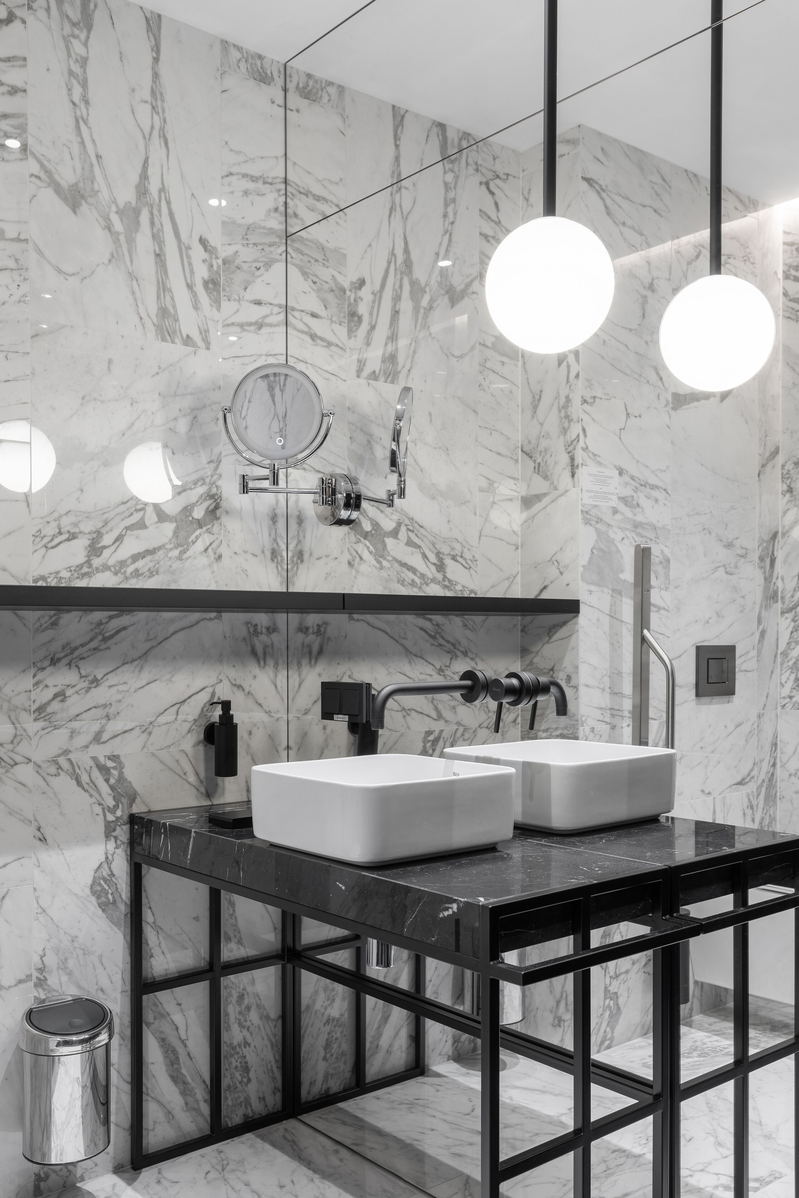 Un baño moderno con paredes de mármol, un mueble de tocador negro, dos lavabos, un espejo y lámparas colgantes.