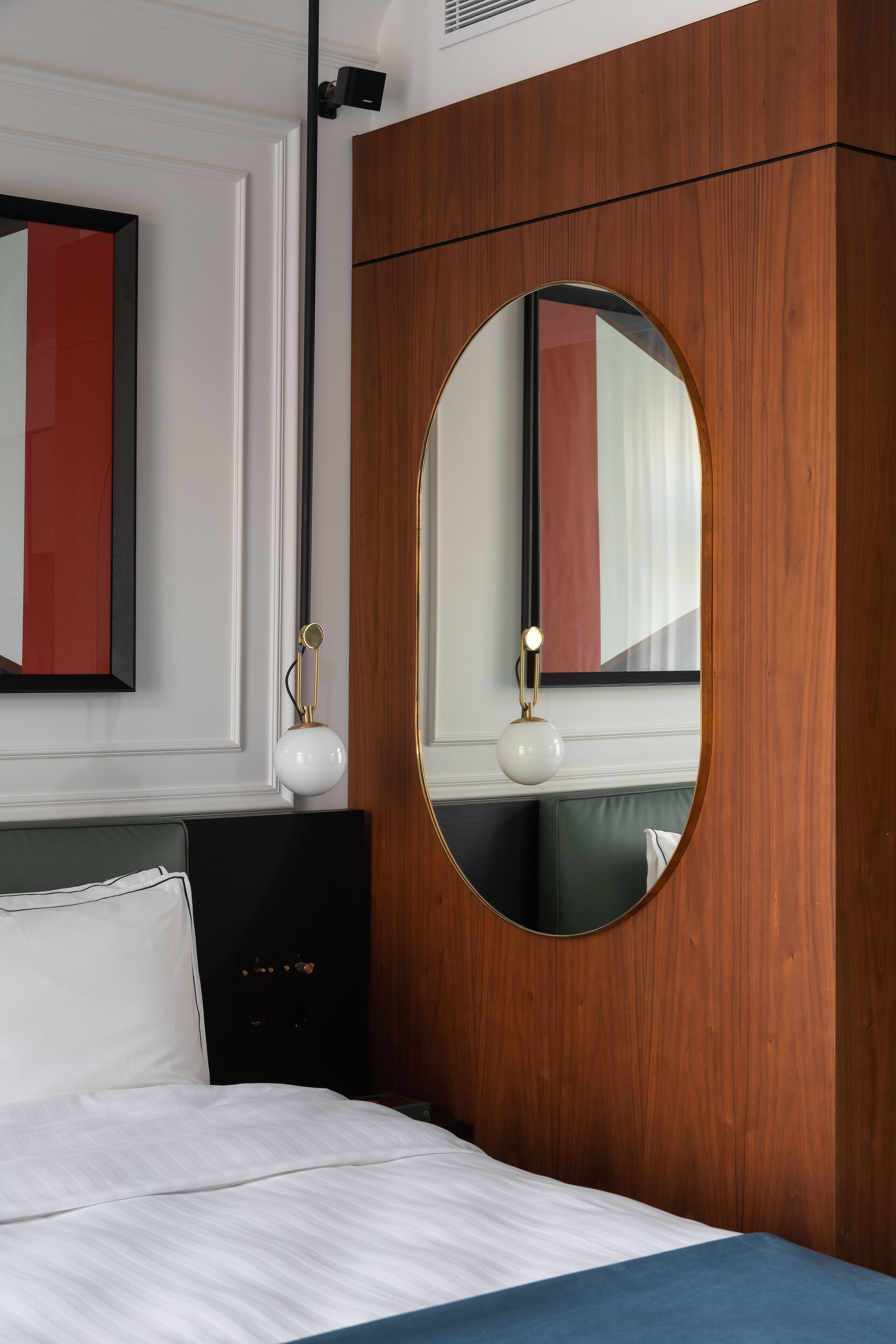 Une chambre d’hôtel avec un lit, un miroir rond, une table de chevet et un luminaire fixé au mur.