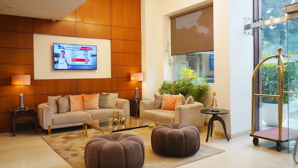 A modern hotel lobby with a large TV screen displaying information, a sofa set, ottomans, a coffee table, and a view of greenery outside. - Alivaa Hotel, Millennium City Center, Sector 29, Gurugram (Gurugram)
