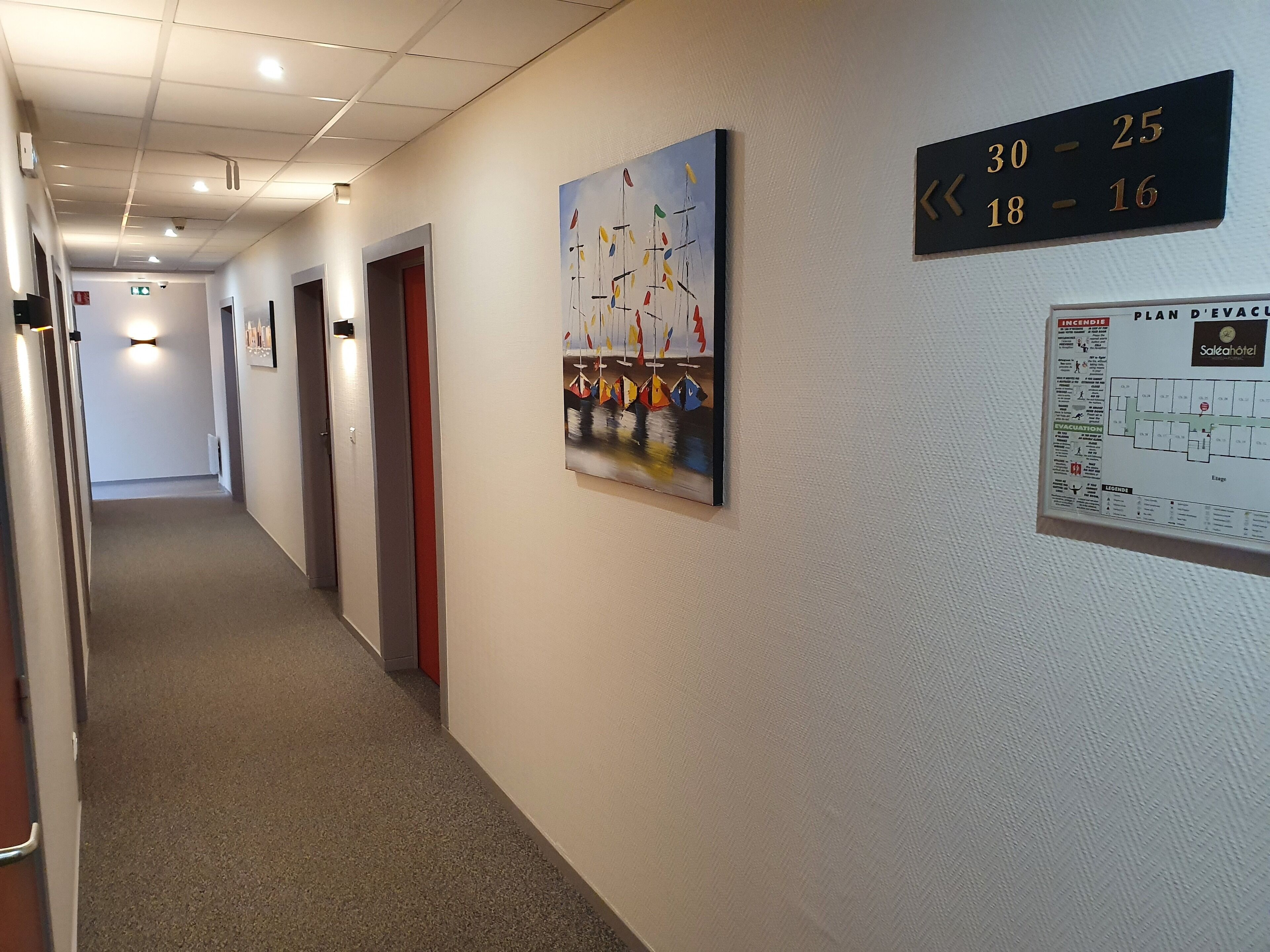A hotel corridor with doors, a painting, and emergency exit signs.
