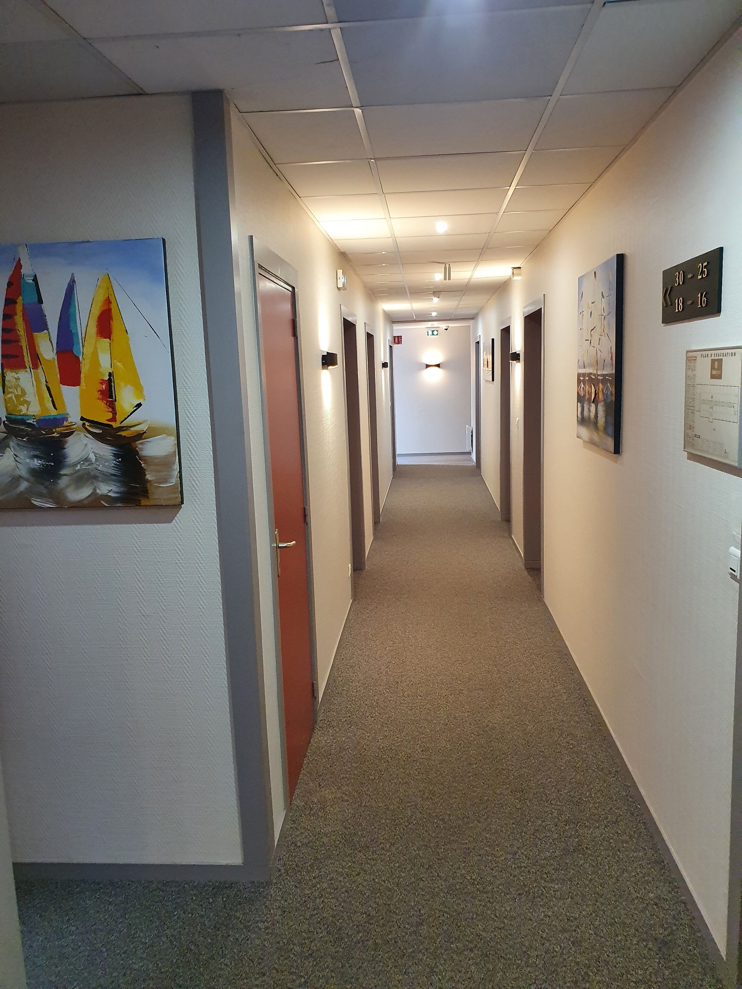 Un couloir d’hôtel avec un sol recouvert de moquette, des portes de chaque côté et des œuvres d’art accrochées aux murs.