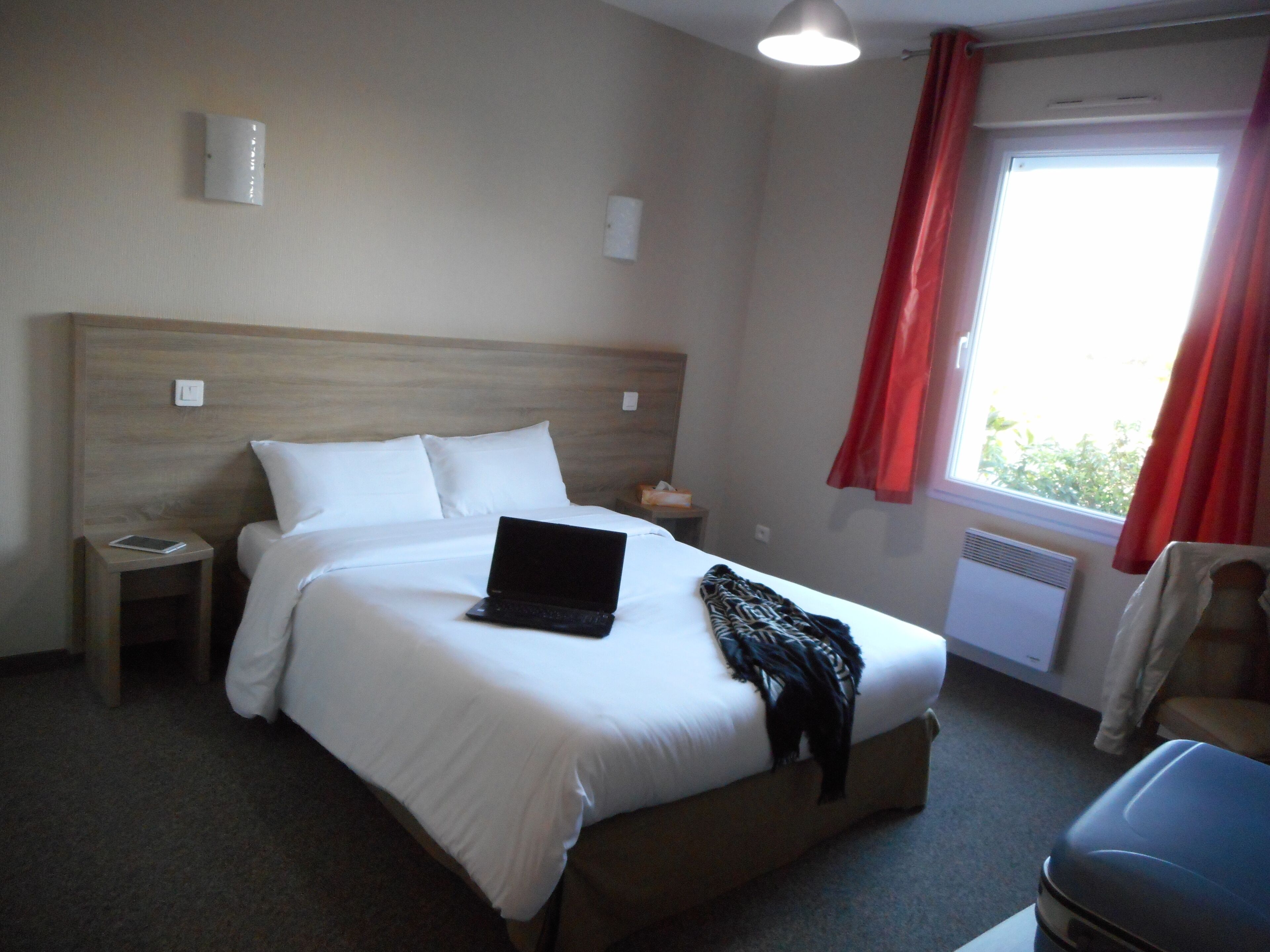 A hotel room with a bed, a laptop on the bed, a nightstand, a chair, a window with red curtains, and a wall-mounted light fixture.