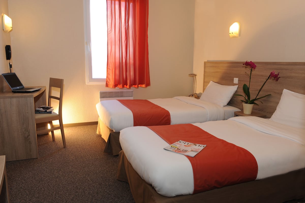 A hotel room with two beds, a desk with a laptop, a chair, a window with red curtains, and a nightstand with a lamp and a potted plant.