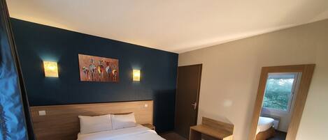 A hotel room with a large bed, bedside tables, a small wooden bench, a mirror, and a window with a view of greenery.