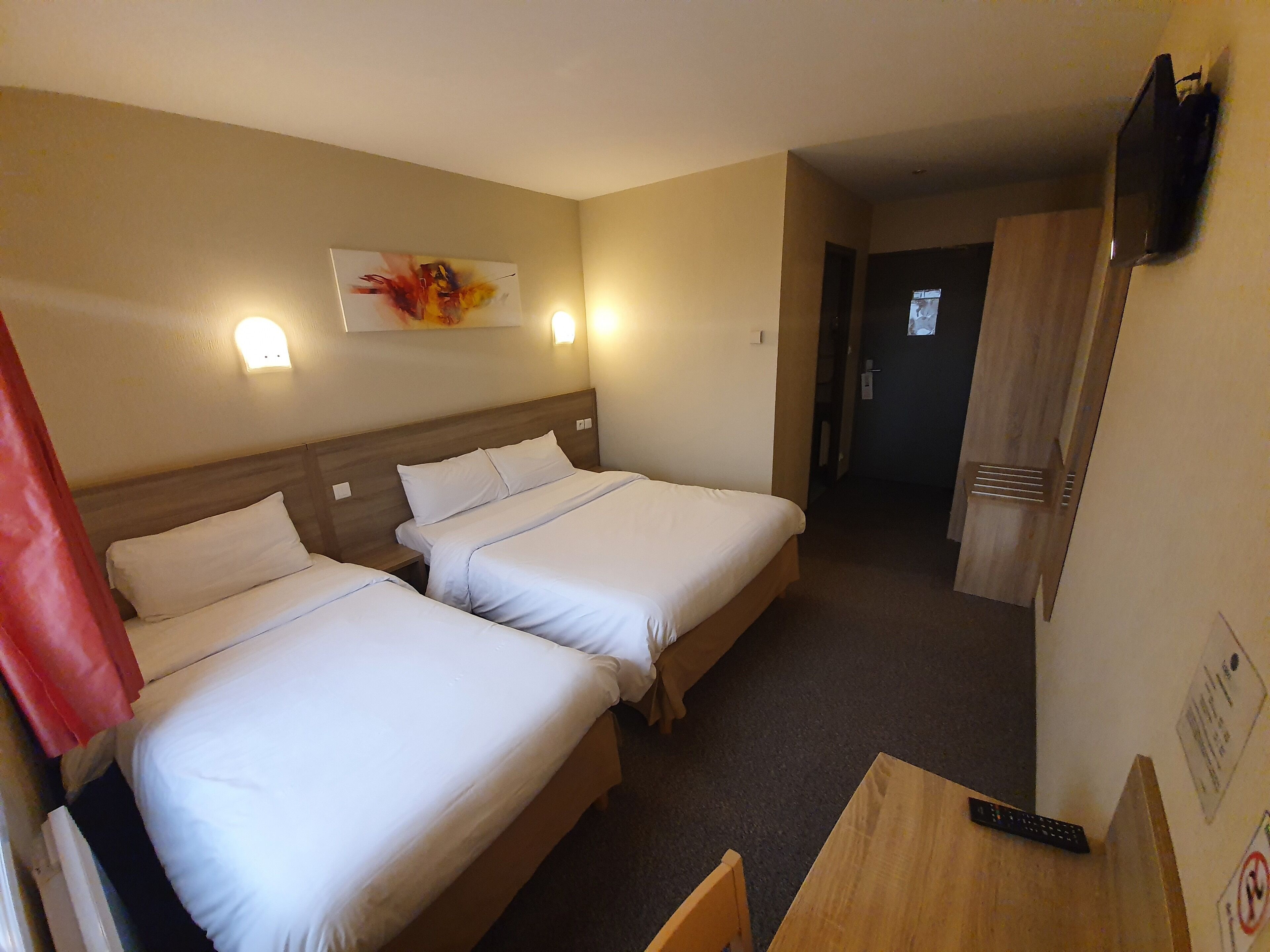 A hotel room with two beds, a wooden headboard, a painting on the wall, a TV mounted on the wall, and a wooden table with a remote control on it.