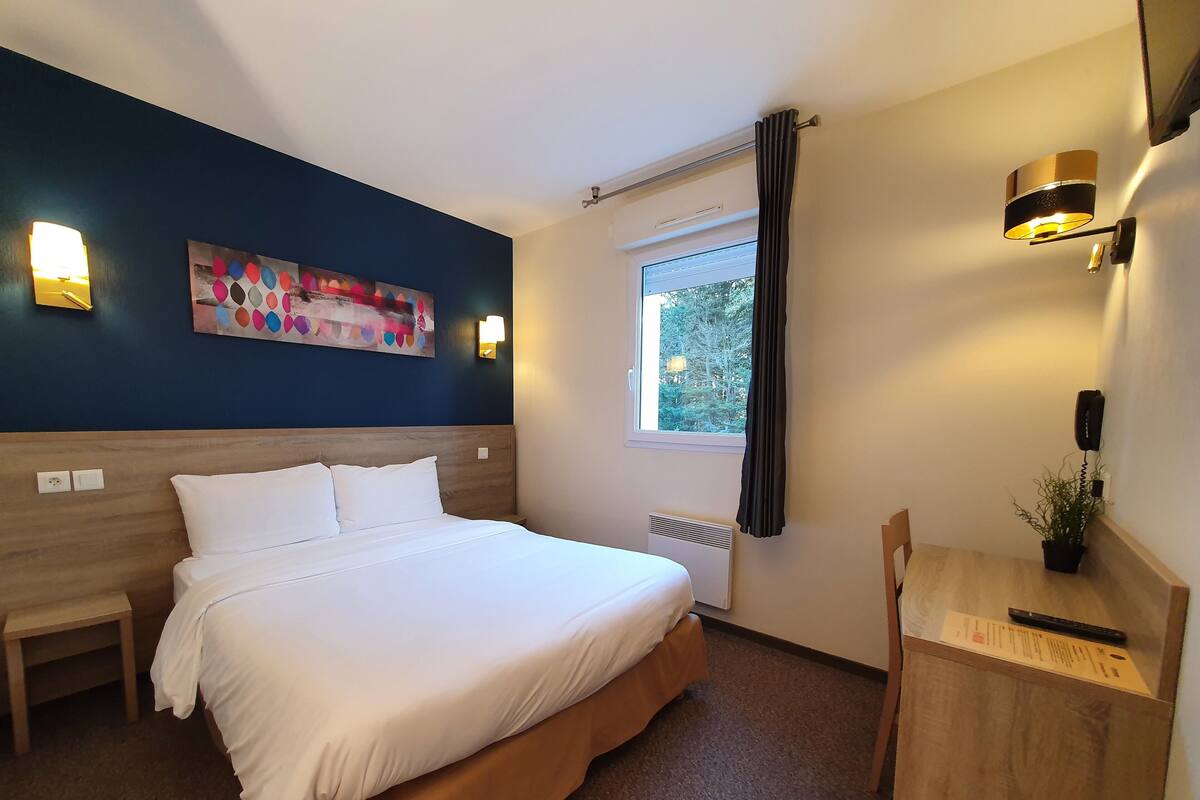 A hotel room with a bed, bedside table, lamp, and a window with curtains.
