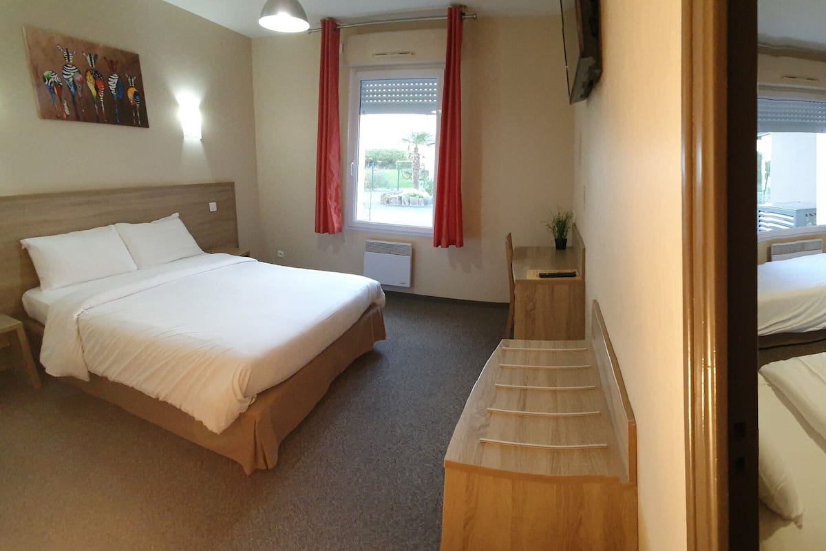 A hotel room with two beds, a small table, a mirror, and a window with curtains.
