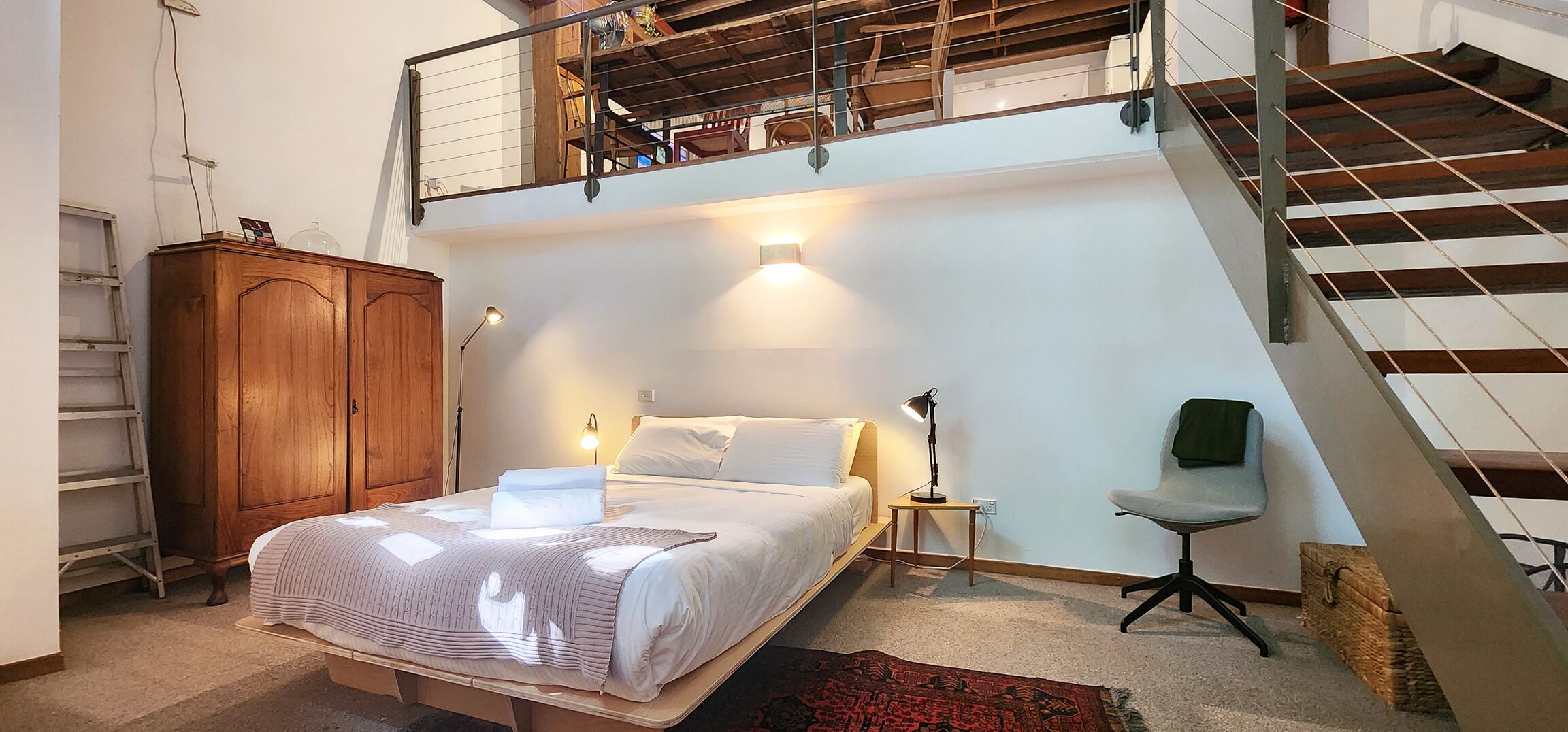 A bedroom with a bed, bedside lamp, chair, wooden wardrobe, and a staircase.