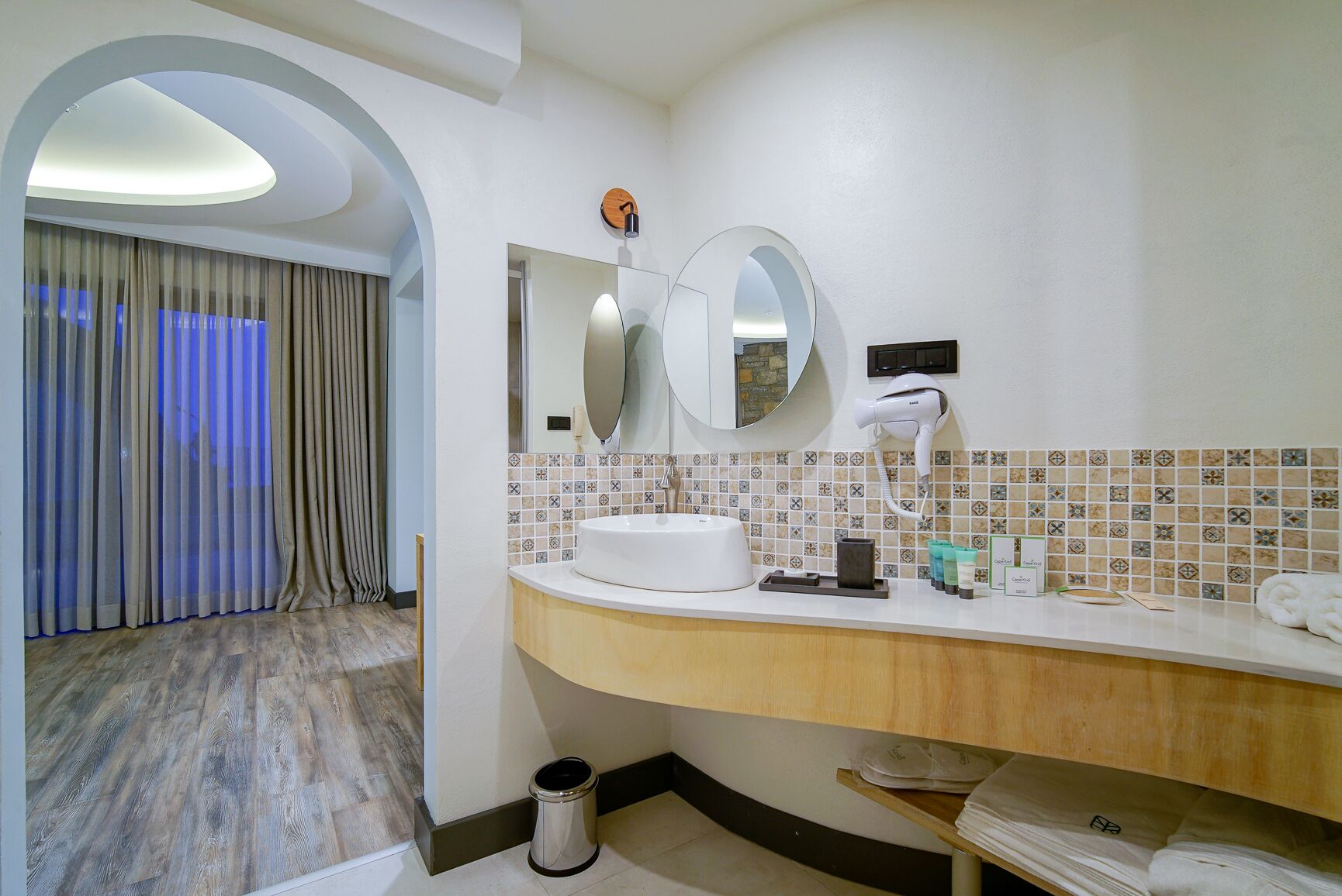A hotel bathroom with a round mirror, a sink, a hairdryer, and a towel rack.