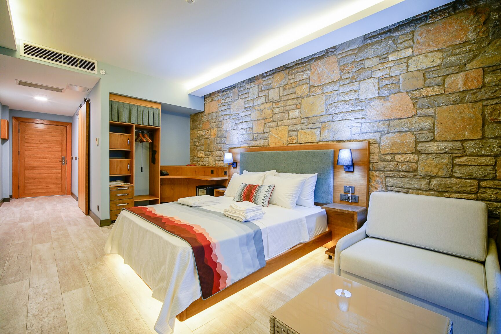 A hotel room with a stone wall, a bed with a headboard, a bedside table, a chair, a wardrobe, and a desk.