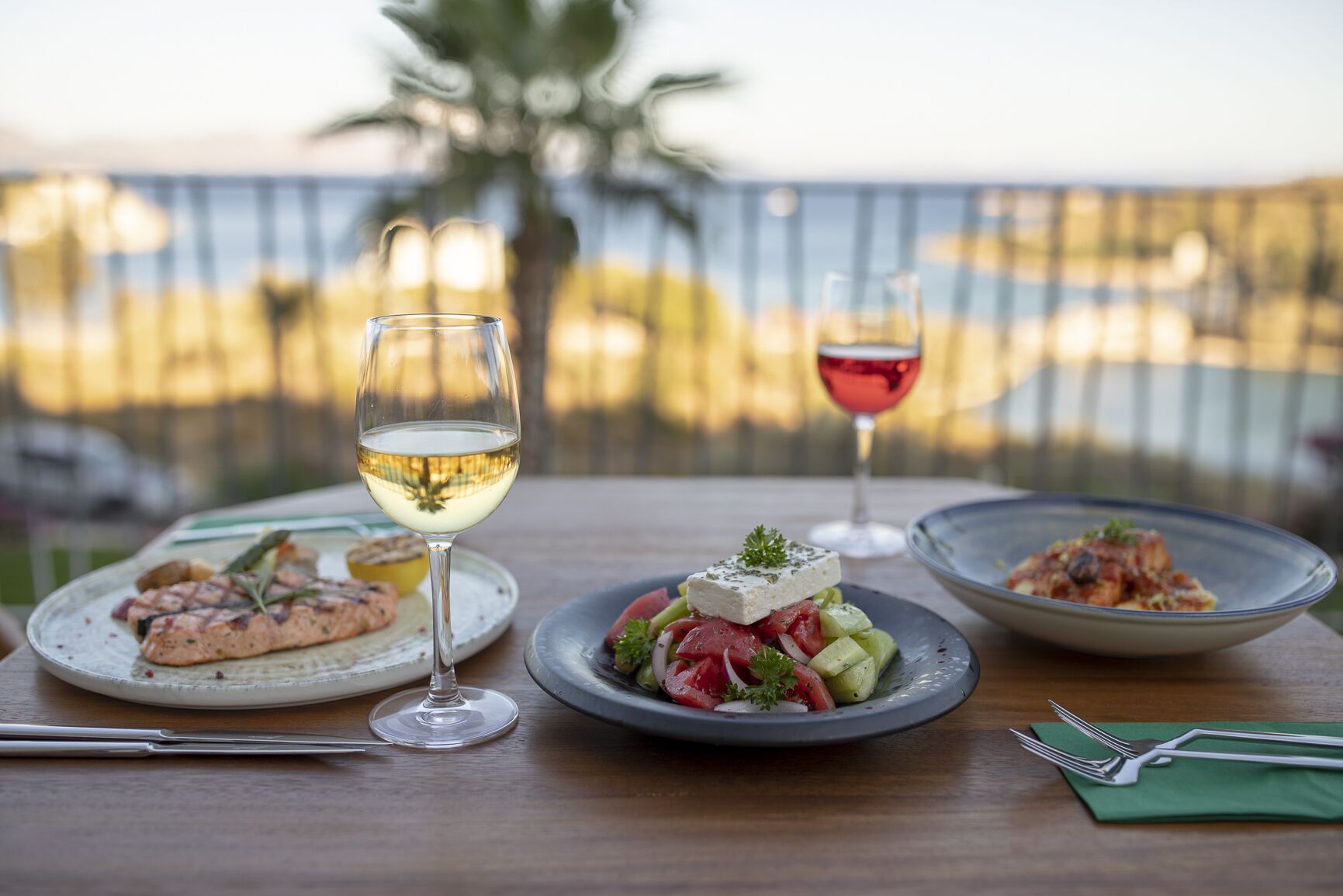 Une table dressée avec trois assiettes de nourriture, deux verres de vin et des couverts, le tout posé sur une table en bois avec une vue sur la mer en arrière-plan.
