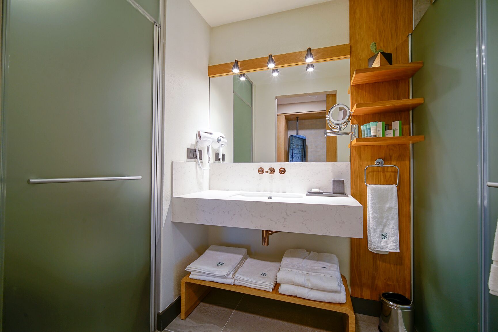 A bathroom with a white sink, a mirror above it, and a wooden shelf with toiletries. There are towels stacked on a wooden bench.