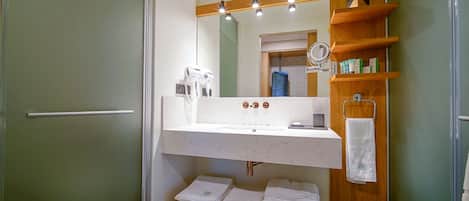 A bathroom with a white sink, a mirror above it, and a wooden shelf with toiletries. There are towels stacked on a wooden bench.