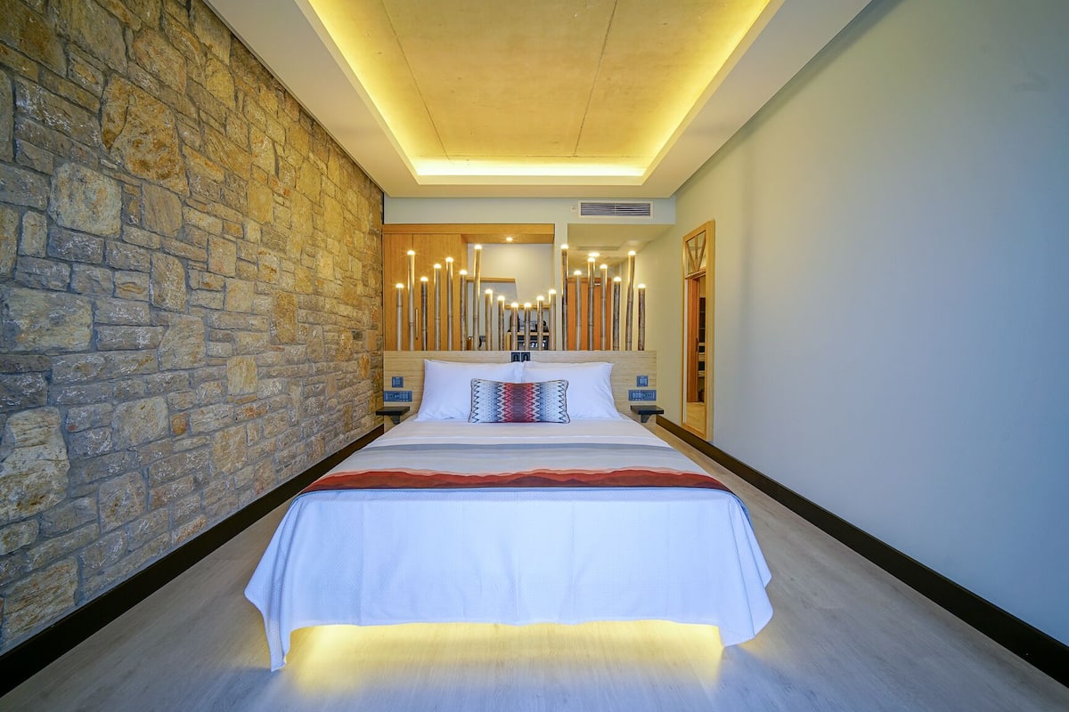 Habitación de hotel moderna con pared de acento de piedra, una cama grande con colcha blanca y cabecero de madera.